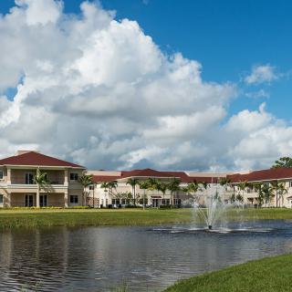 A picturesque view of buildings with red-tiled roofs reflected in a serene lake, featuring a central fountain under a partly cloudy sky.