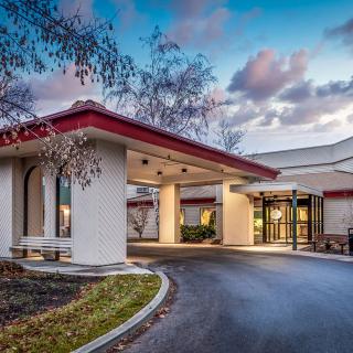 A modern hotel entrance with a driveway and landscaped surroundings during sunset.