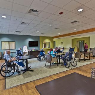 A common area in a nursing home with residents sitting in wheelchairs and chairs, surrounded by warm-toned decor.