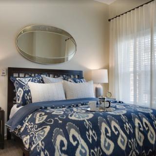 A cozy bedroom featuring a dark wooden bed with patterned blue and white bedding, adorned with pillows. An oval mirror hangs above the bed, and a lamp sits on the bedside table, softly illuminating the room which is brightened by natural light from the wi