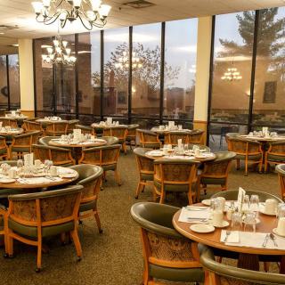 An empty dining room with neatly set tables and chairs, featuring large windows and chandeliers.