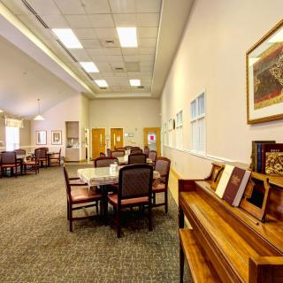 A spacious and well-lit room featuring a dining area with tables, chairs, a piano, and artwork on the walls.