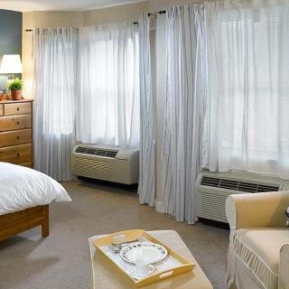 A cozy bedroom with large windows, sheer curtains, a comfortable chair, and a wooden dresser.