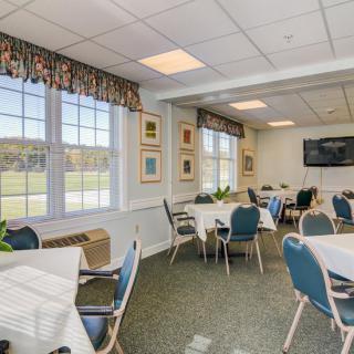 A well-lit dining room with tables and chairs, large windows, and a television on the wall.
