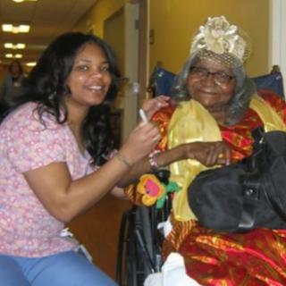 A healthcare worker smiling next to an elderly woman in a wheelchair, dressed for a celebration.