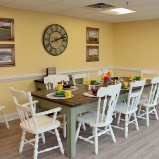 A brightly decorated dining room with a large table and chairs, featuring a wall clock and framed artwork.