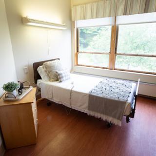 A cozy and well-lit bedroom featuring a single bed, nightstand, and large windows.