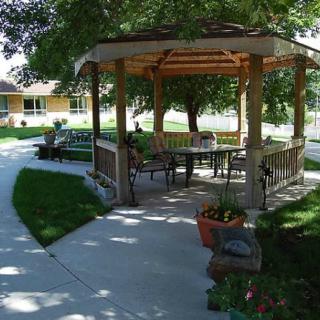 A serene outdoor setting featuring a gazebo with seating surrounded by lush greenery and trees, alongside a winding concrete walkway.