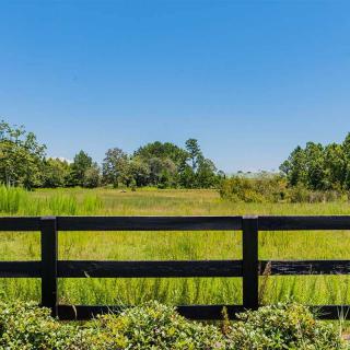 A scenic view of a grassy field bordered by a black wooden fence, with trees in the distance under a clear blue sky.
