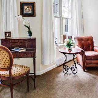 A cozy living room with elegant furniture, featuring a chair, small desk, and side table near a window.
