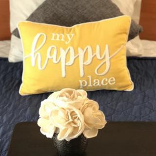 A cozy bedroom scene with a yellow pillow that reads 'my happy place' and a vase of flowers.