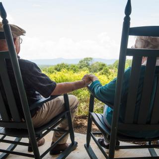 An elderly couple sits on rocking chairs, holding hands and enjoying a scenic view from their porch.