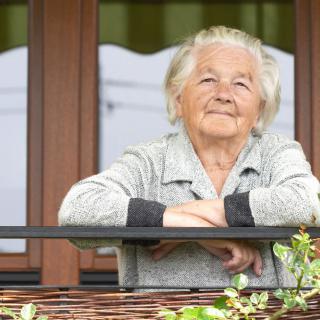 An elderly woman looking thoughtfully into the distance while standing on a balcony.
