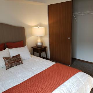 A cozy bedroom featuring a neatly made bed with orange accents, a bedside lamp, and an open closet.