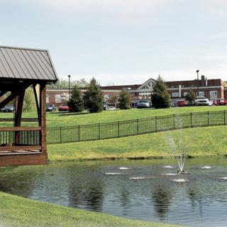 A serene park featuring a pond with a fountain, a wooden pavilion, and lush green grass with buildings in the background.