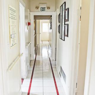 A bright hallway with tiled flooring, lined with doors and framed pictures, leading to an exit.