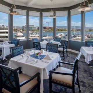 A waterfront restaurant with elegantly set tables, offering a stunning view of the marina through large windows.