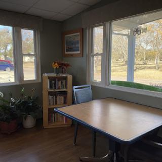 A cozy corner of a room featuring a table, bookshelf, and natural light streaming through large windows.