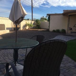 A backyard patio with a glass table, chairs, and an umbrella, set on a paved area under a sunny sky.