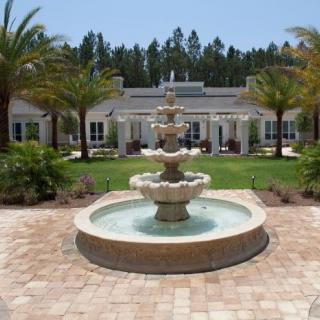 A beautifully landscaped courtyard featuring a large tiered fountain, surrounded by palm trees and a building in the background.