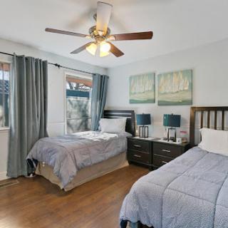 A cozy and well-lit bedroom featuring two twin beds, a ceiling fan, and soft blue-gray decor.