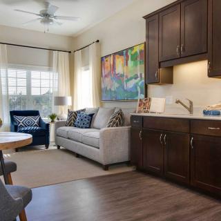 A cozy living space with a small kitchen area, featuring a sofa, armchair, and artwork on the wall.
