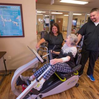 An elderly person engaged in exercise with the assistance of healthcare professionals on fitness equipment indoors.