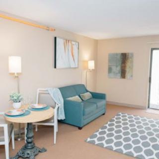 A cozy living room with a teal sofa, artwork on the walls, a small dining table, and a patterned rug.