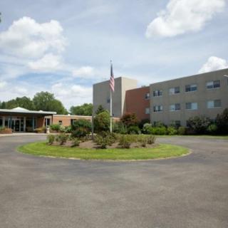 A hospital building with a circular driveway, landscaped garden, and flagpole under a partly cloudy sky.