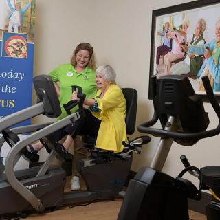 An elderly woman receives assistance while using exercise equipment in a fitness room.