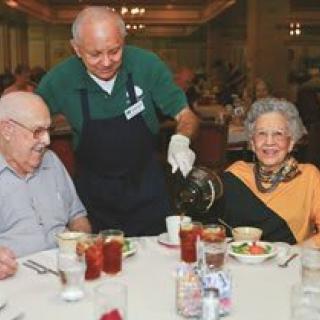 An elderly couple enjoys a meal at a restaurant while being served by a waiter.