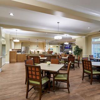 A well-lit dining area featuring multiple tables and chairs, with a view of the kitchen in the background.