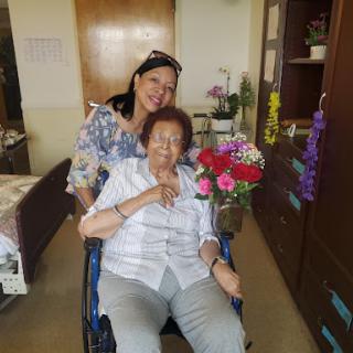A joyful moment shared between a caregiver and an elderly woman in a wheelchair surrounded by beautiful flowers.