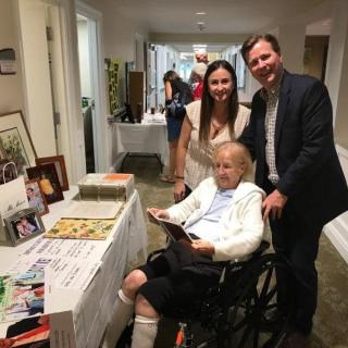 A group of people smiling together at an indoor event, with one person seated in a wheelchair next to a table displaying various items.
