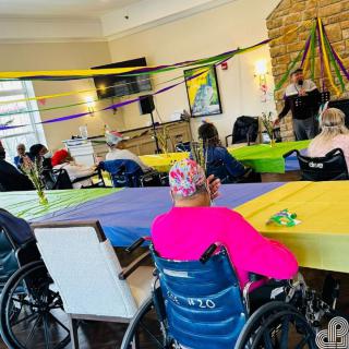 A gathering at a nursing home with seniors in wheelchairs attending an indoor event, decorated with colorful streamers.