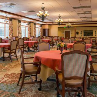A well-lit dining room with several round tables covered in red tablecloths and surrounded by upholstered chairs.