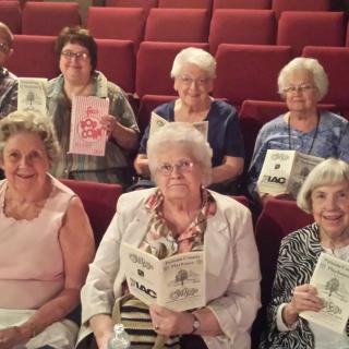 A group of elderly women sitting in an auditorium, holding books.