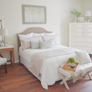 A cozy, minimalist bedroom featuring a neatly made bed with light-colored bedding and decorative pillows, a dresser, and soft lighting.