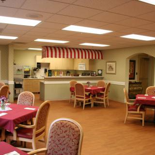A cozy dining area featuring neatly arranged tables with red tablecloths and a small open kitchen in the background.