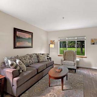 A cozy living room with a sofa, armchair, and coffee table, featuring a large window and decorative elements like a painting and rug.