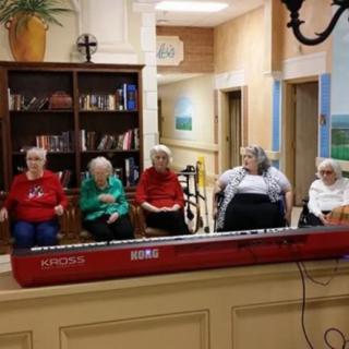 A group of elderly people sitting in a room with a keyboard and bookshelf.