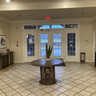 A spacious lobby area with tiled flooring, featuring a central table adorned with a plant and large windows providing natural light.