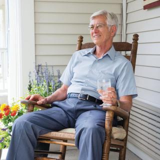 An elderly man relaxing in a rocking chair on a porch, holding a glass and surrounded by flowers.