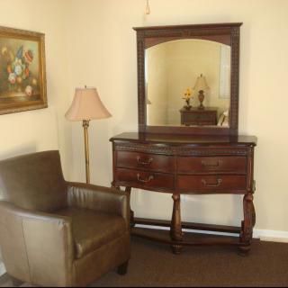A cozy room corner featuring a vintage dresser with a mirror, an armchair, a floor lamp, and a wall painting.