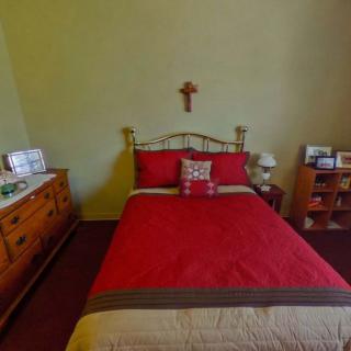 A cozy bedroom featuring a red bedspread with a wooden cross on the wall above the bed, flanked by a dresser and nightstand.