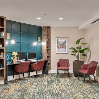A cozy office workspace featuring two computer stations with red chairs, a patterned carpet, decorative wall paneling, and indoor plants for a welcoming atmosphere.