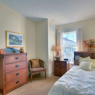 A cozy bedroom with a wooden dresser, armchair, and a neatly made bed near a window with soft lighting.