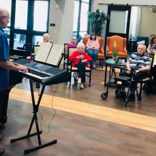 A musician plays the keyboard for an audience of elderly individuals in a cozy indoor setting.