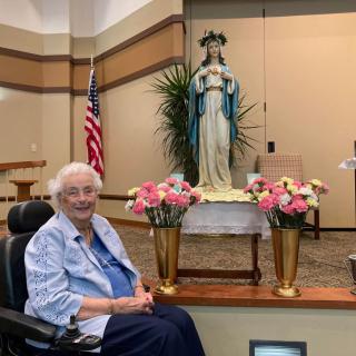 An elderly woman in a wheelchair sits near a religious statue adorned with flowers, with an American flag in the background.