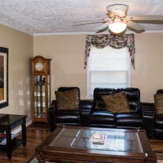A cozy living room with a leather sofa, glass-top coffee table, grandfather clock, and ceiling fan.